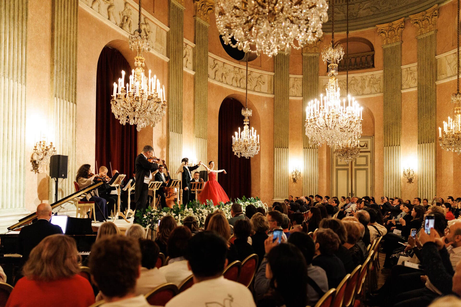 A soprano in a red gown sings beneath crystal chandeliers in the Baroque hall of Palais Auersperg