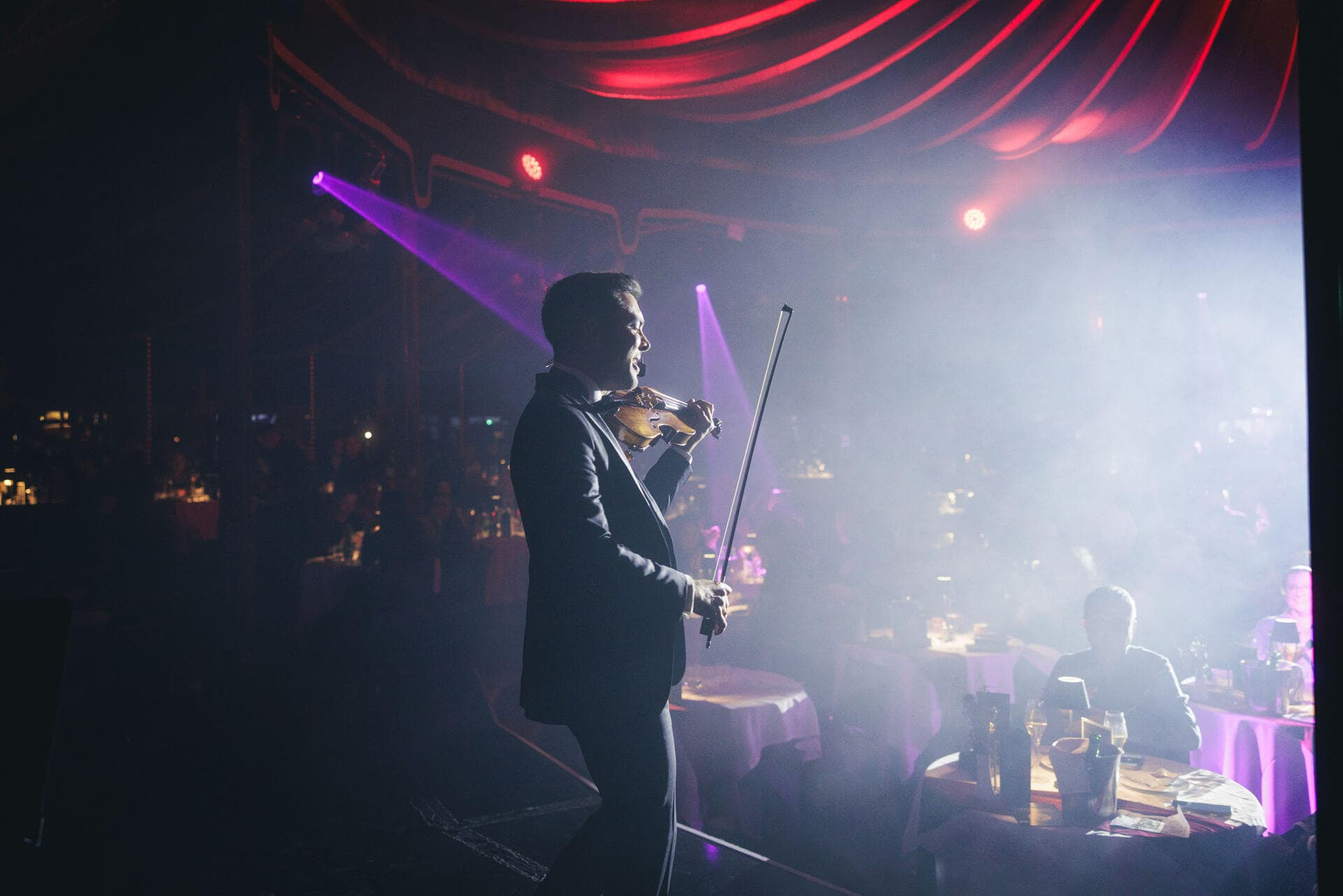 A violinist performing on a dark stage under warm red and violet lights at the Strauss Dinner Show